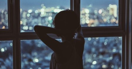 Person blickt nachts aus dem Fenster auf eine erleuchtete Stadt – Symbol für Schlaflosigkeit und Schlafkrise